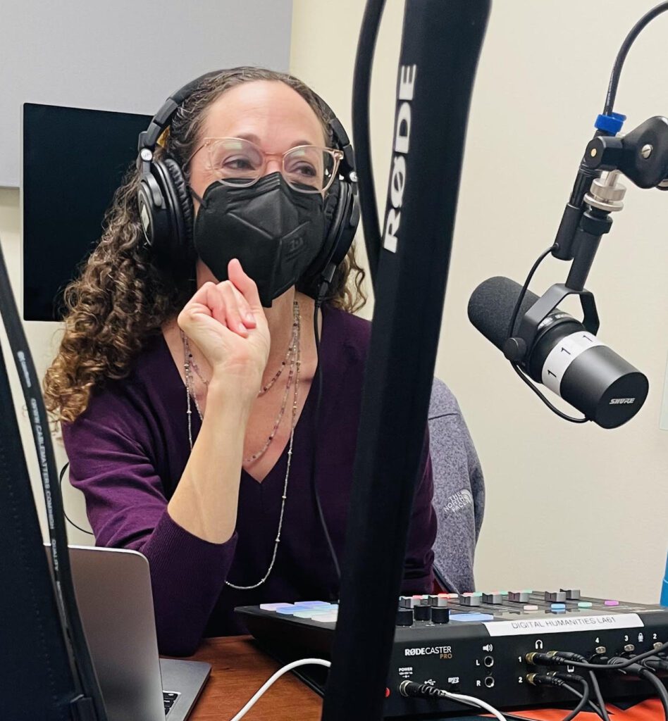 White woman wearing black mask and headphones sits at a sound board in front of a mic