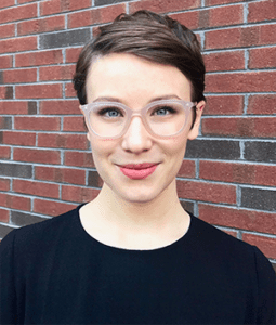 White woman wearing glasses and a black sweater in front of a brick wall